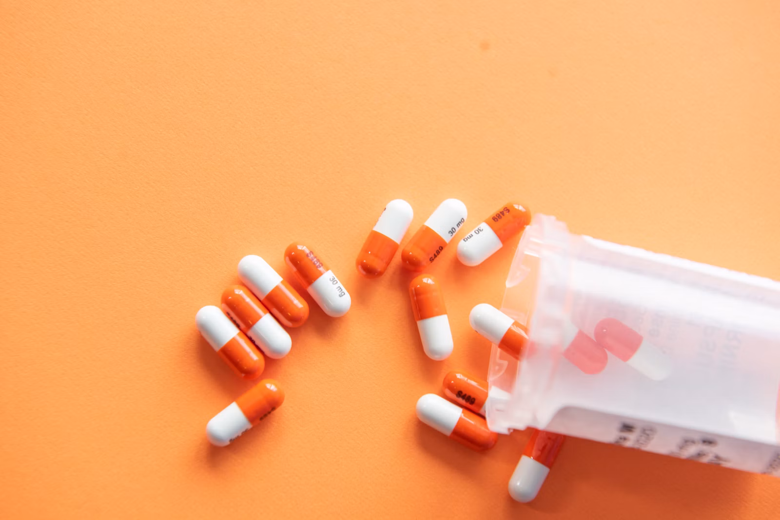 Orange and white capsules spilled from a clear prescription bottle on an orange surface